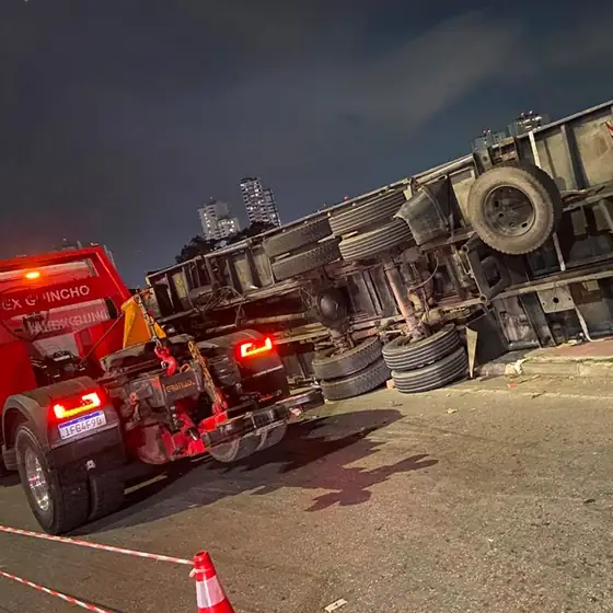 Caminhão caçamba tombado à noite com guincho lança engatado e área sinalizada